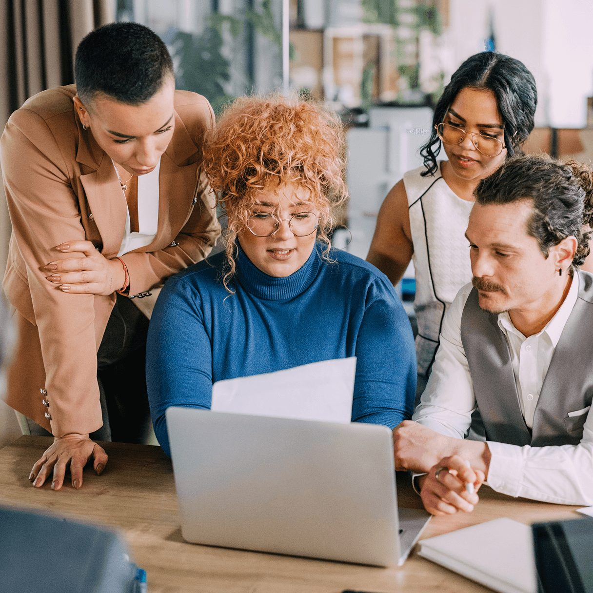 Quatro colegas se reúnem em volta de uma mesa, olhando atentamente para a tela do laptop e para os documentos durante uma reunião colaborativa.