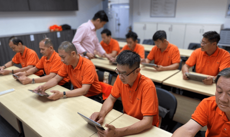 Adult men in orange shirts use tablets in a classroom with an instructor.