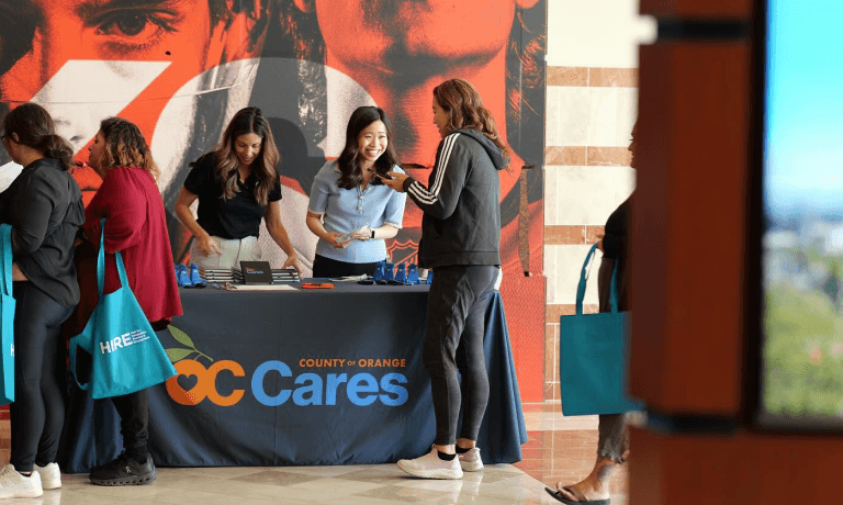 Women interact at a "County of Orange OC Cares" event booth, with some carrying "HIRE" tote bags.
