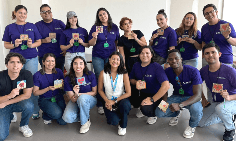 A diverse group of smiling individuals, many wearing purple A.M.S. shirts, holding up small decorated cards.