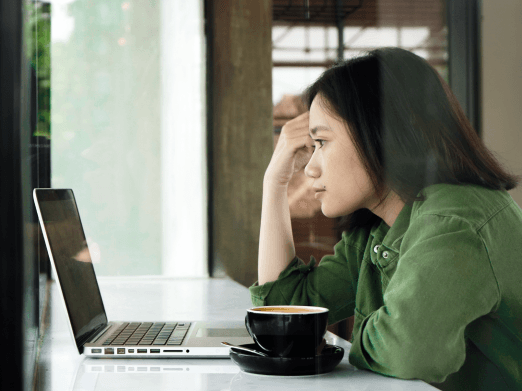 Young woman in a green shirt sitting at a cafe table with a laptop and coffee, looking thoughtfully out a window.