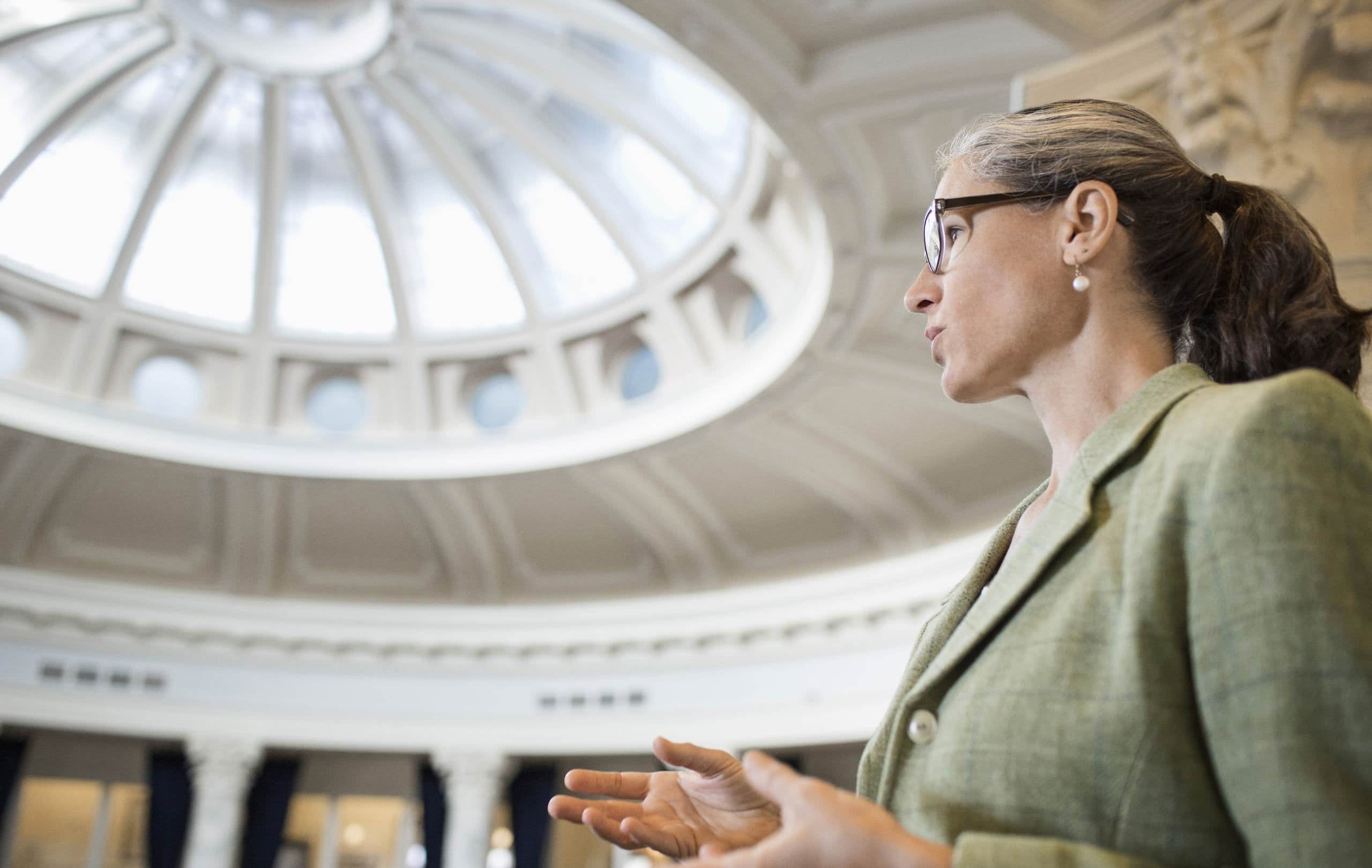 Woman in glasses and a green blazer speaking in a domed hall.
