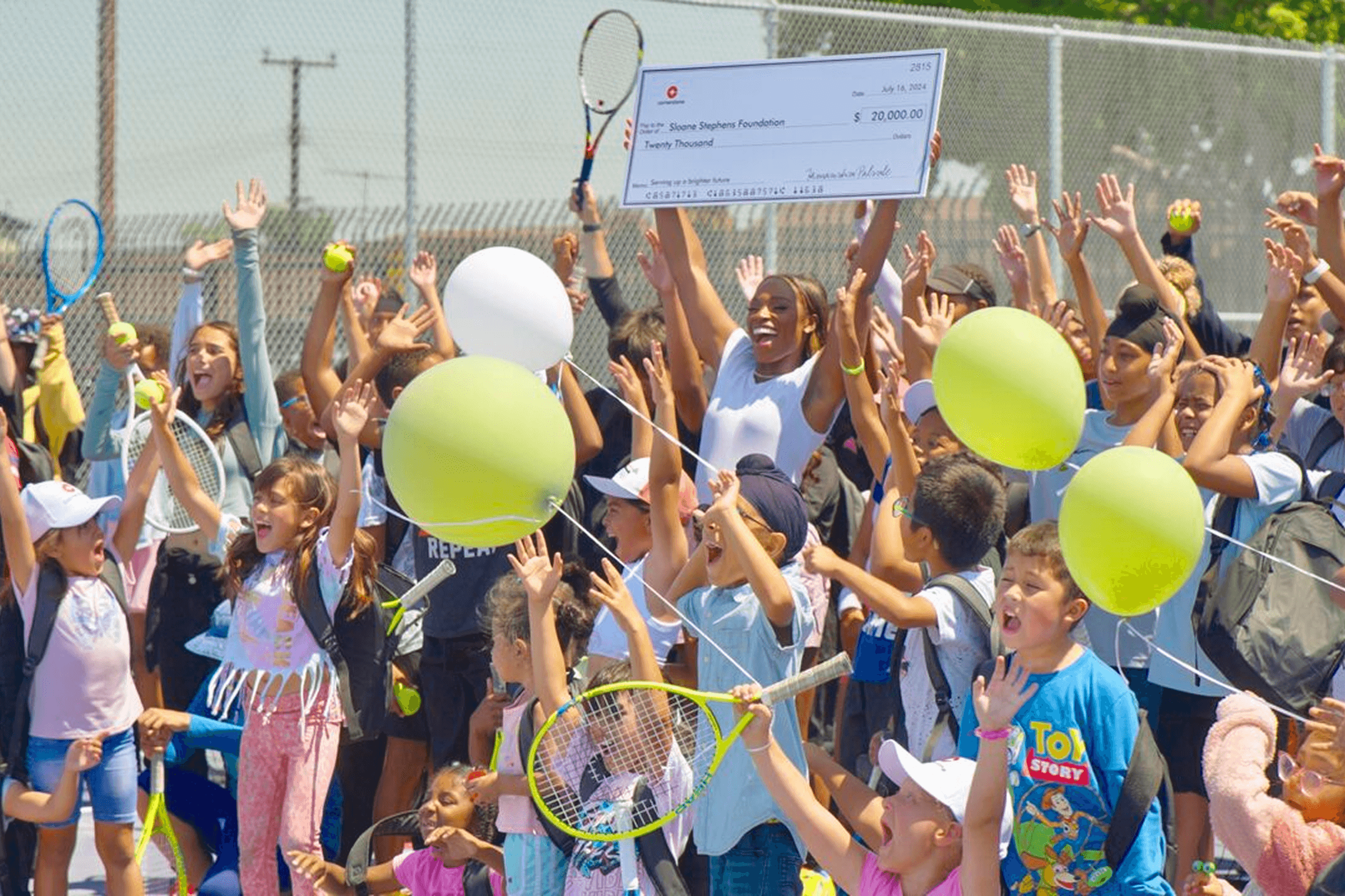 Tennis player Sloane Stephens holds a $20,000 check for her foundation, surrounded by excited children holding tennis rackets and balls on a court.
